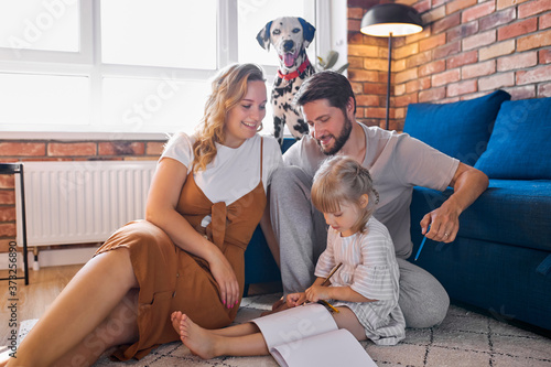 friendly family love spedning time with daughter at home, young parents sit with girl on the floor, she is painting, drawing photo