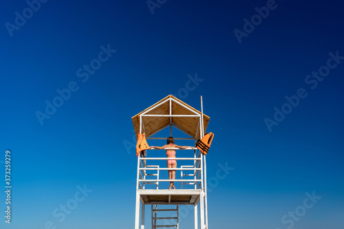 rear view. teenage girl stands on lifeguard tower on beach against cloudless sky.