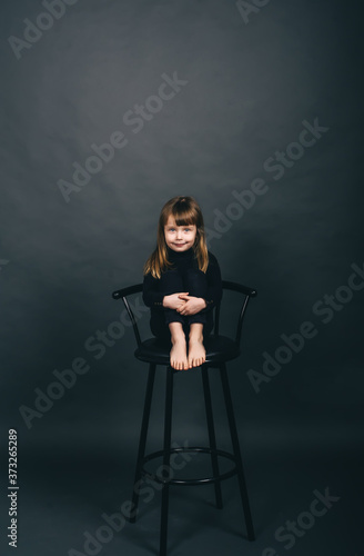 Beatiful little girl with long hair in a black clothes and barefoot sits on a chair in the studio