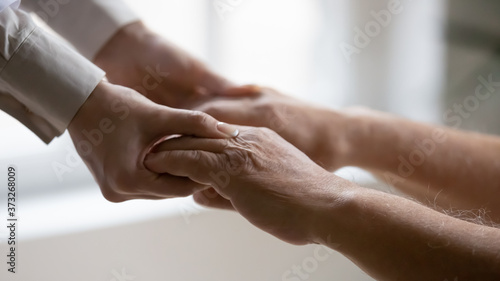 Close up side view young female doctor nurse holding wrinkled hands of elderly mature patient, helping standing up. Old 80s man involved in rehabilitation process with professional therapist indoors.