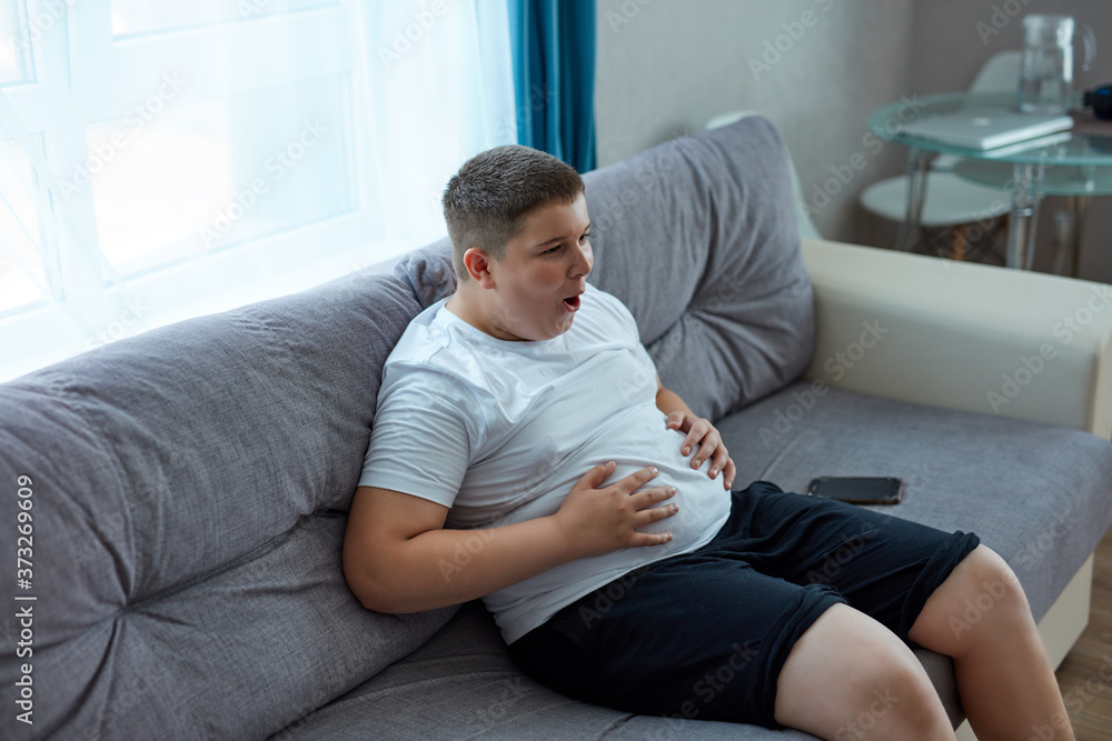 fat teenager boy has stomach ache after junk food, sit on sofa holding belly, suffer from pain