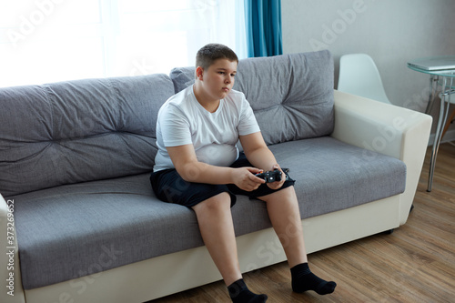 fat caucasian teenager boy enjoys video games, overweight boy sits on sofa holding console in hands, concentrated on game, look at screen