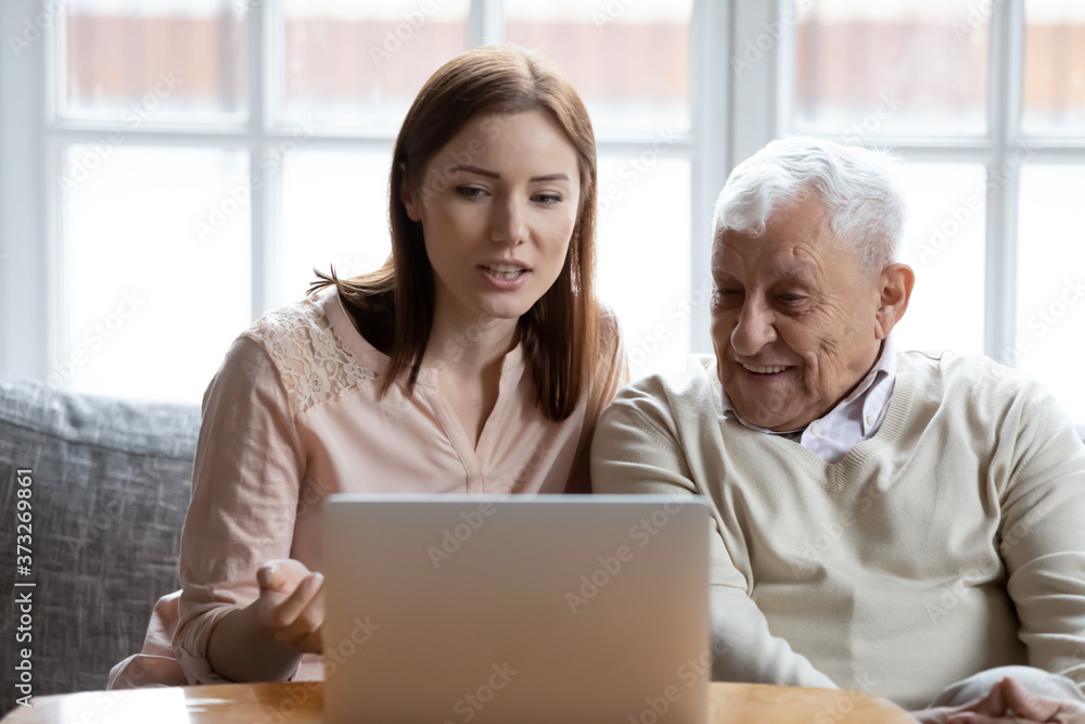 Front view happy elderly 70s grey haired man learning using computer software with caring grown up daughter, sitting on couch in living room, retired old people and modern technology concept. Front view happy elderly 70s grey haired man learning using computer software with caring grown up daughter, sitting on couch in living room, retired old people and modern technology concept.