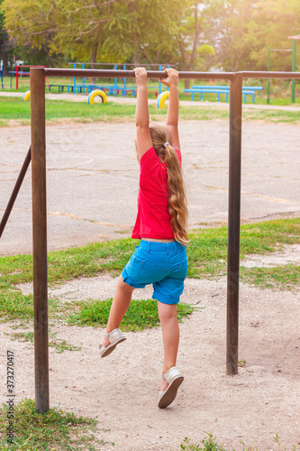 Girl hanging on horizontal bar. Child doing motion exercises and playing on playground outdoor. Healthy leisure sport activity on fresh air. Happy childhood. Kids physical training on sports stadium.