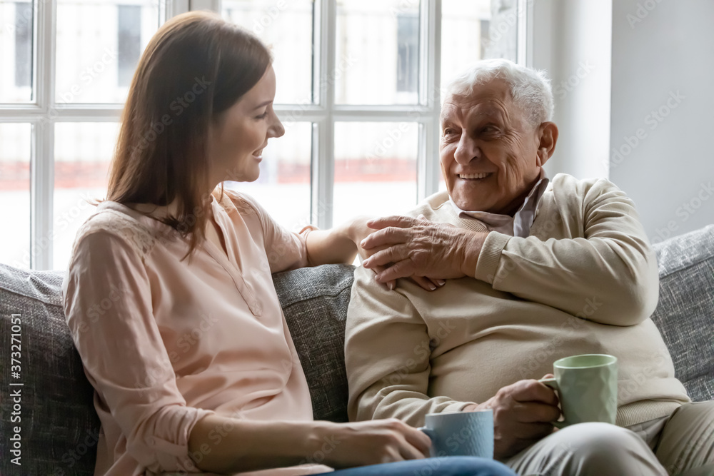 Smiling young woman sitting on sofa with happy older retired 70s father, enjoying pleasant conversation with cup of coffee tea together in living room, mature parents and grown children communication. Smiling young woman sitting on sofa with happy older retired 70s father, enjoying pleasant conversation with cup of coffee tea together in living room, mature parents and grown children communication.
