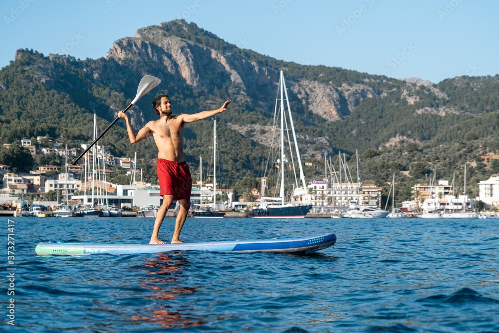 Young man with a red boardshorts practising stand up paddle surf SUP at Port of Soller (Serra de Tramuntana, Mallorca, Spain) with the mountains at the background