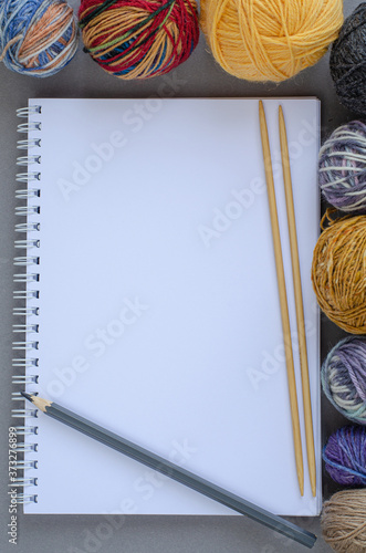 Background with multicolored yarn balls and notebook with pencil and knitting bamboo needles on gray background. Craft, handmade, hobby concept. Copy space, top view