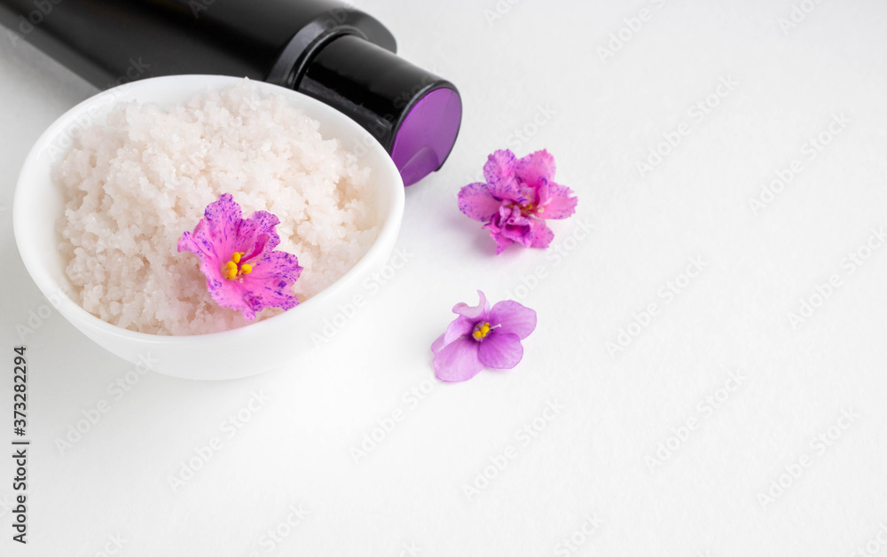 An oval white bowl with pink salt stands on a white background next to it are a black bottle and violet flowers