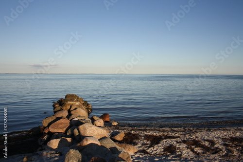 Steinstrand mit einem Haufen Steine im Vordergrund. Im Hintergrund sieht man dasMeer und den Himmel am Horizont. 