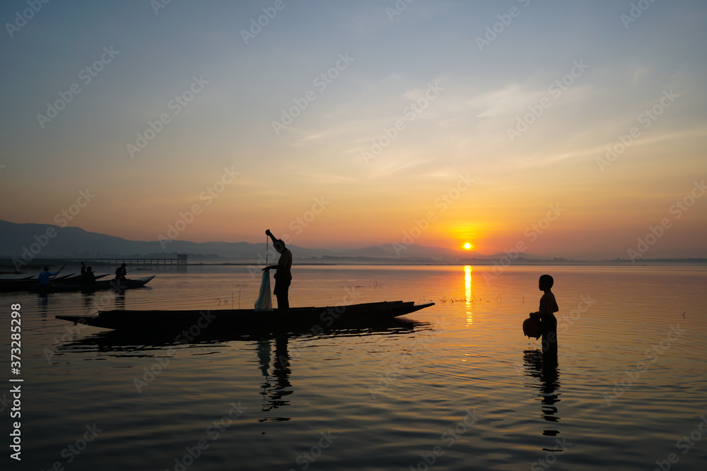 asian fisherman standing on boat using fishing net to catch fish while his son standing and holding jar in the lake at the sunrise