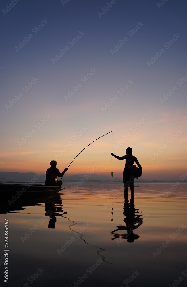 At lake side, asian fisherman sitting on boat while his son standing and  using fishing rod to catch fish at the sunrise