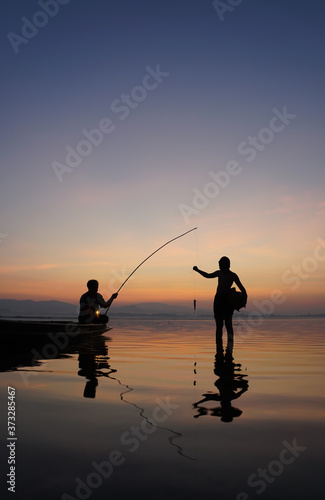 At lake side, asian fisherman sitting on boat while his son standing and using fishing rod to catch fish at the sunrise