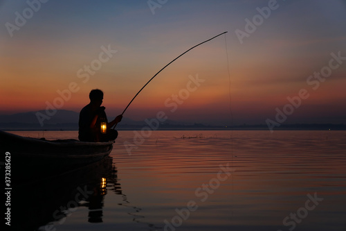 At lake side, asian fisherman sitting on boat and using fishing rod to catch fish at the sunrise © plo