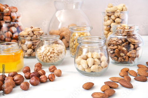 Different nuts (hazelnuts, cashews, pistachios, pine nuts, walnuts, almonds and chickpeas) in glass jars stand on a white background. There is a small jar of honey nearby. Healthy organic food
