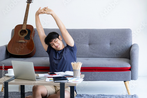 Young Asia man working in the living room are stretch oneself. Sitting on the ground and leaning against the sofa with the guitar. There are laptop, phone tablet a cup off coffee on the table.