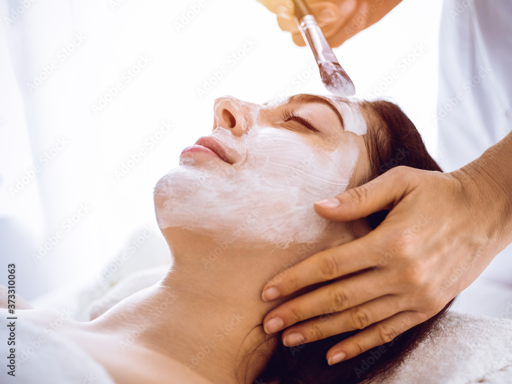 Naklejka premium Beautiful brunette woman enjoying applying cosmetic mask with closed eyes in sunny spa center. Relaxing treatment and medicine concept