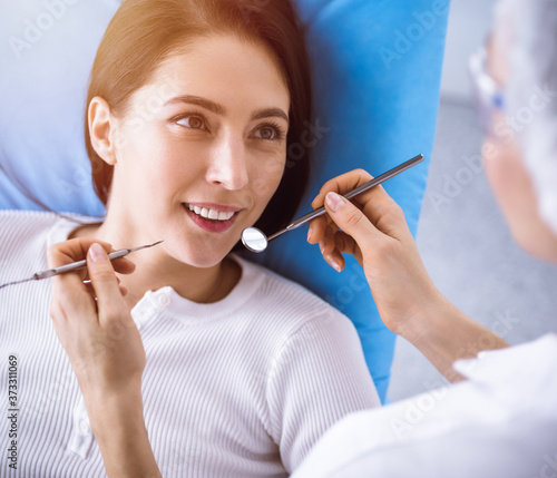 Smiling brunette woman being examined by dentist at sunny dental clinic. Hands of a doctor holding dental instruments near patient s mouth. Healthy teeth and medicine concept
