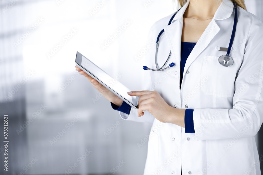 Unknown woman-doctor is holding a tablet computer in her hands, while standing in a clinic. Female physician with a stethoscope in her office, close-up. Perfect medical service in a hospital. Medicine Unknown woman-doctor is holding a tablet computer in her hands, while standing in a clinic. Female physician with a stethoscope in her office, close-up. Perfect medical service in a hospital. Medicine