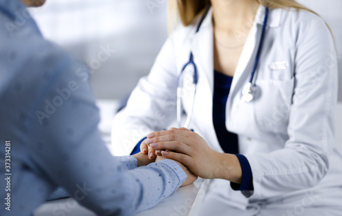 Unknown woman-doctor is holding her patient's hands to reassure the patient, discussing current health examination, while they are sitting together at the desk in the cabinet in a clinic. Female