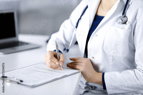 Unknown woman-doctor is prescribing some medication for her patient, using a clipboard, while sitting at the desk in her cabinet. Female physician with a stethoscope, close up. Perfect medical service
