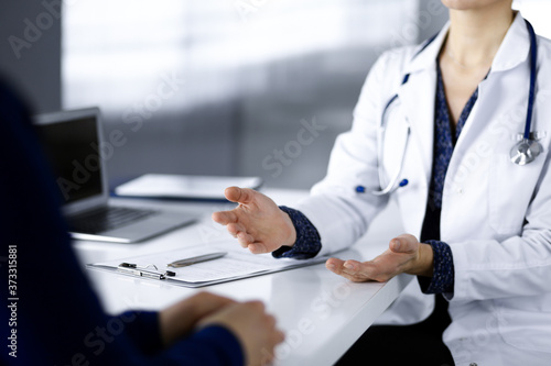 Unknown woman-doctor is talking to her patient about her diagnosis, while sitting together at the desk in the cabinet in a clinic. Physician at working place, close-up. Perfect medical service in a