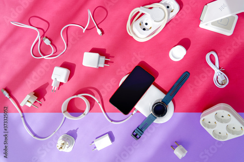 The concept of wireless charging of mobile gadgets. The smartphone and smartwatch are on the wireless charging pad. There are various adapters, cables and chargers nearby. Colored paper background.