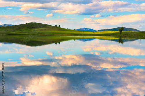 Green hills  blue sky with clouds and their reflections in the lake at sunset. Beautiful summer landscape. South Ural  Russia