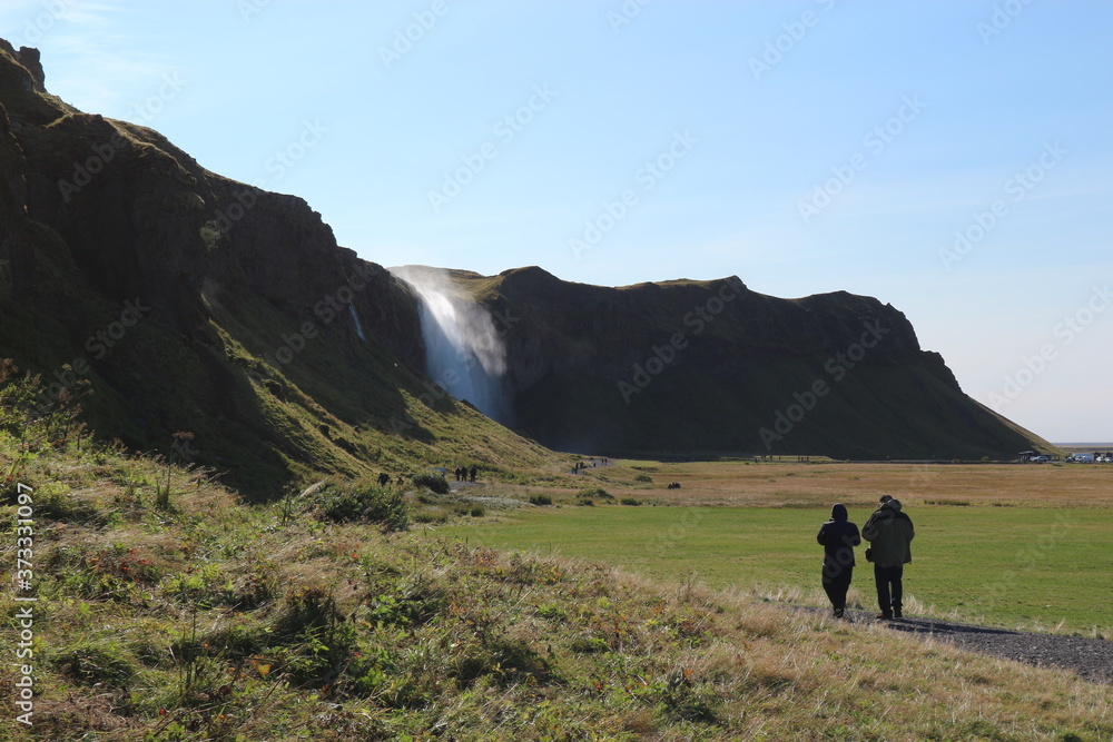 Couple walking together on green lawn with mountain and waterfall in the background. Landscape located in Iceland