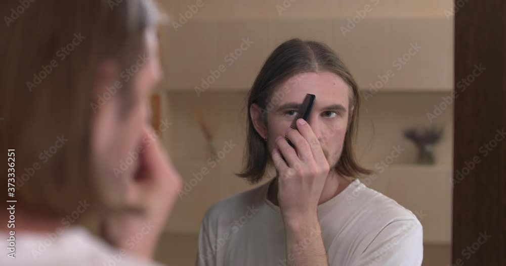 Young man looks at himself in the mirror and styling eyebrows with a comb Young man looks at himself in the mirror and styling eyebrows with a comb