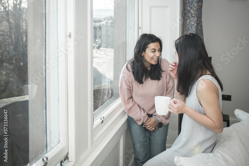 A British-Bangladeshi woman wearing jeans and a pink top and her Asian friend chat and laugh over a cup of tea while standing next to a window in a flat in Edinburgh  Scotland  United Kingdom