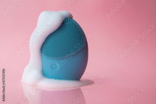 Silicone brush for washing face in foam. Blue facial cleansing brush on pink background. Beauty and skincare concept. Brush for face lifting, anti-aging wrinkles and massage