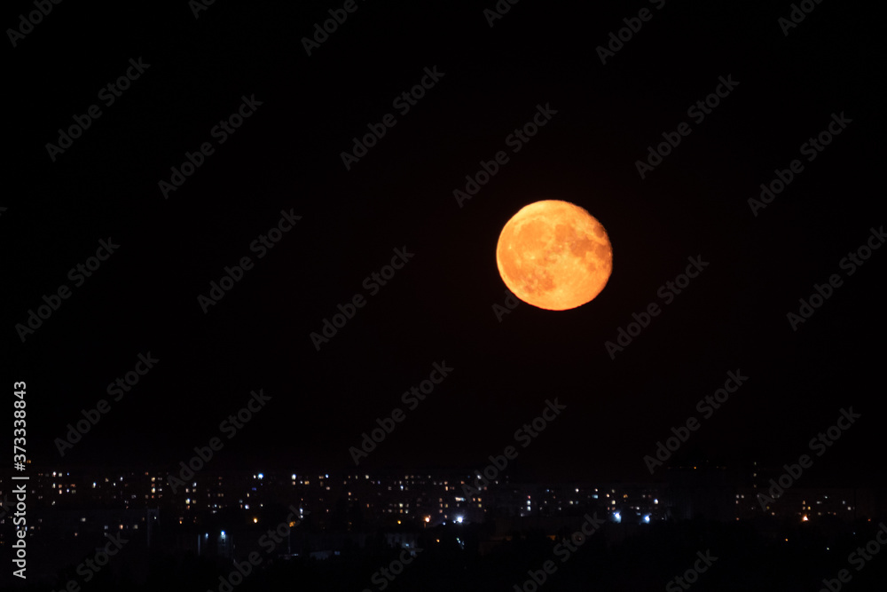 Fototapeta premium Big full red orange moon rising up on dark night sky above city lights. Mystic nighttime black sky with large glowing moon