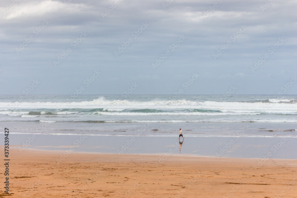 Lonely man walk on seashore on cloudy day. Huge wave on the beach with tourist. Panoramic seascape. Autumn on the coast, Spain. Holiday illustration. Surf sport concept. Seaside nature. Loneliness.