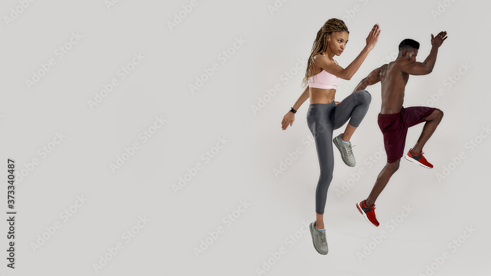 Full length shot of young muscular african american man and sportive mixed race woman looking focused while jumping, exercising isolated over grey background. Sports and workout concept