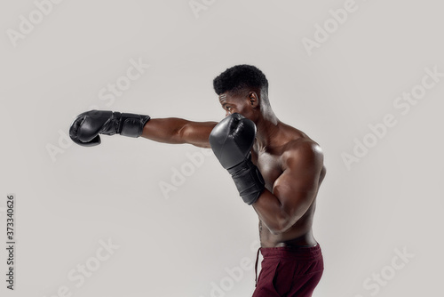 Portrait of young muscular african american male boxer looking aside, wearing boxing gloves, punching, standing isolated over grey background. Sports, workout, bodybuilding concept