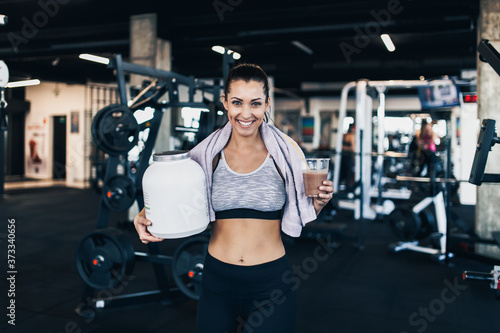 Young attractive woman after successful workout in modern fitness gym holding big white protein jar and drinks shake with drinking straw in other hand.