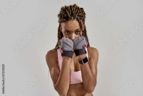 Portrait of young sportive mixed race woman in sportswear looking at camera, wearing fitness gloves while posing isolated over grey background. Healthy lifestyle and sports concept