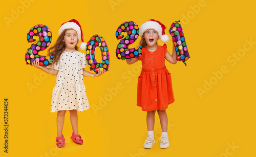 Cheerful children in Santa hats holding colorfull numbers 2021 on yellow background. Happy New Year party with balloons