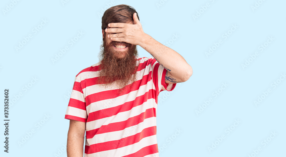 Handsome young red head man with long beard wearing casual clothes smiling and laughing with hand on face covering eyes for surprise. blind concept. Handsome young red head man with long beard wearing casual clothes smiling and laughing with hand on face covering eyes for surprise. blind concept.