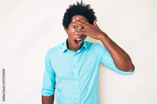 Handsome african american man with afro hair wearing casual clothes peeking in shock covering face and eyes with hand  looking through fingers with embarrassed expression.