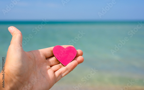 A man s hand holds a pink heart on a blue sea background. Concept for travel and vacation by the sea