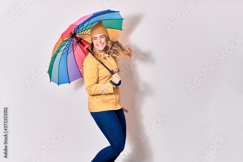 Young beautiful woman wearing winter clothes smiling happy. Jumping with smile on face holding colorful umbrella over isolated white background