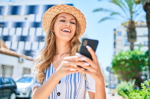 Young beautiful blonde woman on vacation wearing summer hat smiling happy. Standing with smile on face using smartphone at street of city.