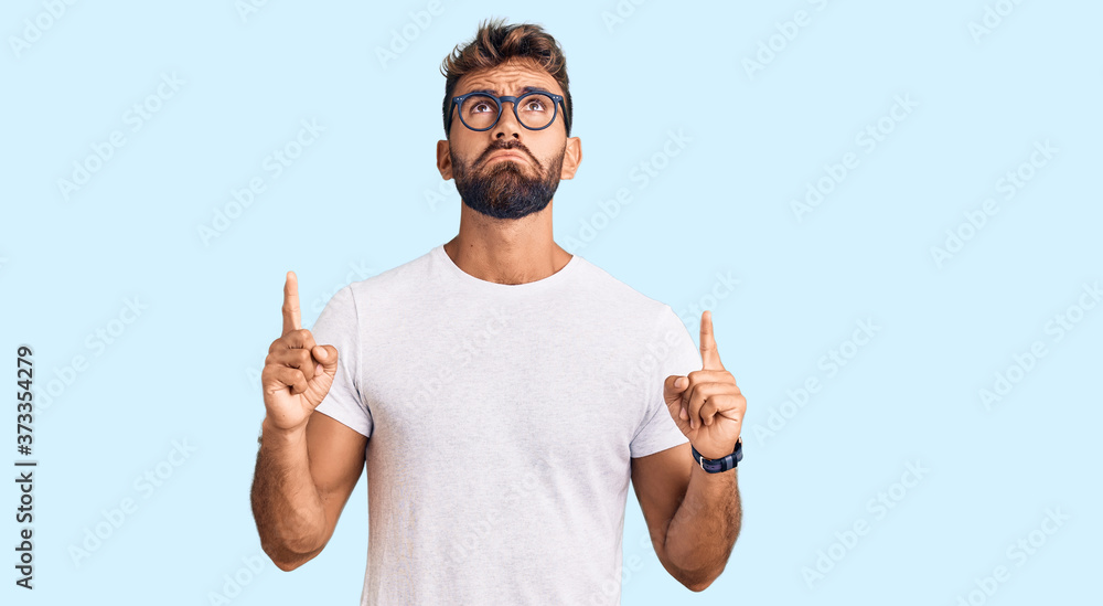 Young hispanic man wearing casual clothes and glasses pointing up looking sad and upset, indicating direction with fingers, unhappy and depressed. Young hispanic man wearing casual clothes and glasses pointing up looking sad and upset, indicating direction with fingers, unhappy and depressed.