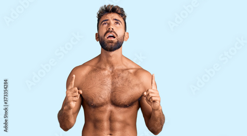 Young hispanic man wearing swimwear shirtless amazed and surprised looking up and pointing with fingers and raised arms.
