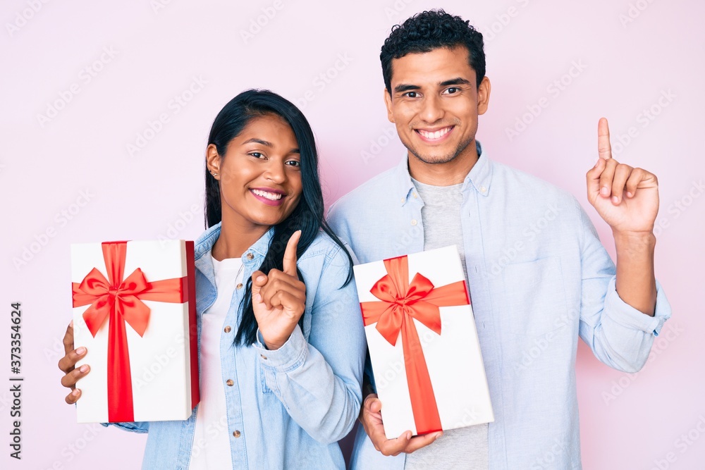 Beautiful latin young couple holding gift for anniversary celebration surprised with an idea or question pointing finger with happy face, number one Beautiful latin young couple holding gift for anniversary celebration surprised with an idea or question pointing finger with happy face, number one