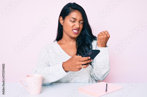 Beautiful latin young woman with long hair using smartphone sitting on the table annoyed and frustrated shouting with anger, yelling crazy with anger and hand raised