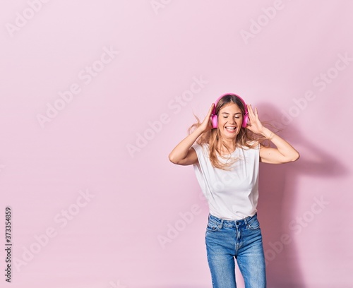 Young beautiful blonde woman smiling happy. Jumping with smile on face listening to music using pink headphones over isolated background