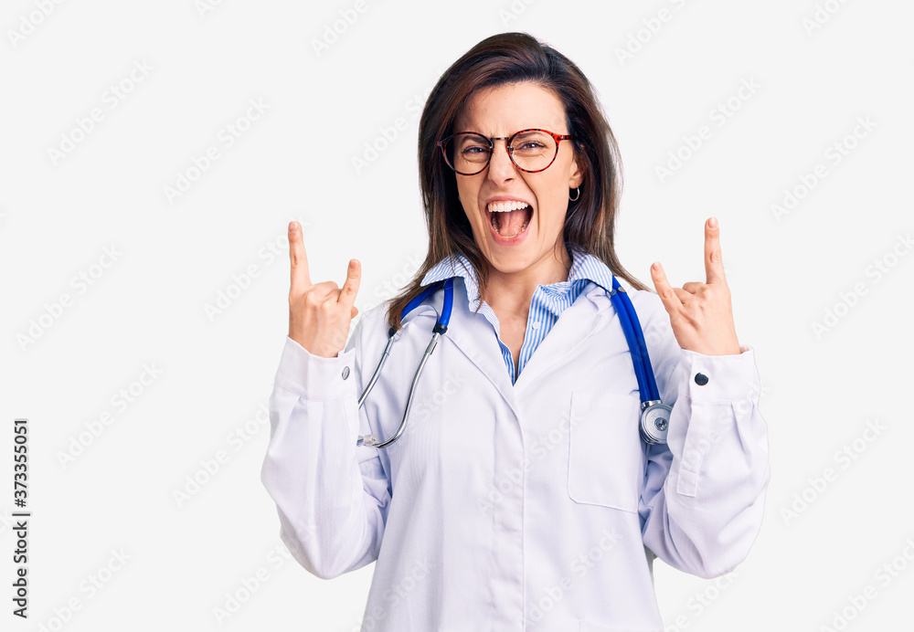 Young beautiful woman wearing doctor stethoscope and glasses shouting with crazy expression doing rock symbol with hands up. music star. heavy concept. Young beautiful woman wearing doctor stethoscope and glasses shouting with crazy expression doing rock symbol with hands up. music star. heavy concept.