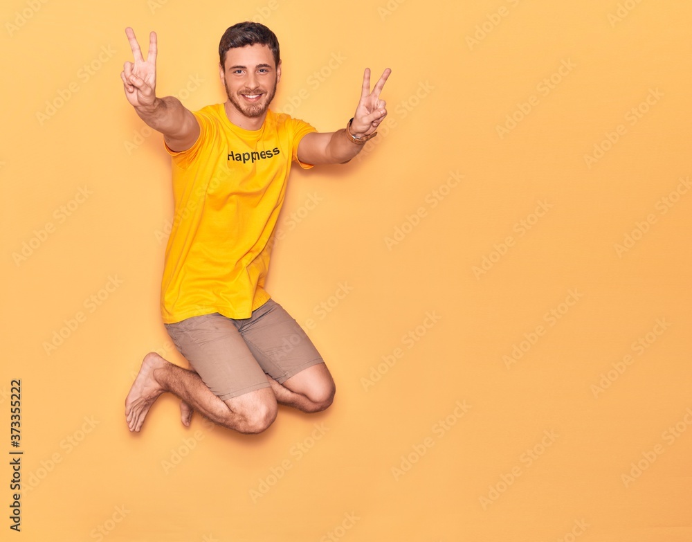Young hispanic man wearing t shirt with happiness word message smiling happy. Jumping with smile on face doing victory sign over isolated yellow background Young hispanic man wearing t shirt with happiness word message smiling happy. Jumping with smile on face doing victory sign over isolated yellow background